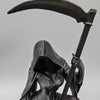 A dark figurine of a female grim reaper wearing a hood and holding a large scythe against a gray background.
