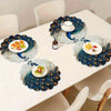 A set of four elegant peacock feather placemats is displayed on a white dining table with food and a teacup.