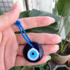 A hand holds a dark blue glass evil eye amulet pendant with a white iris and blue thread against a blurred green plant backgr