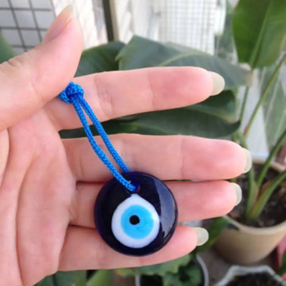 A hand holds a dark blue glass evil eye amulet pendant with a white iris and blue thread against a blurred green plant backgr