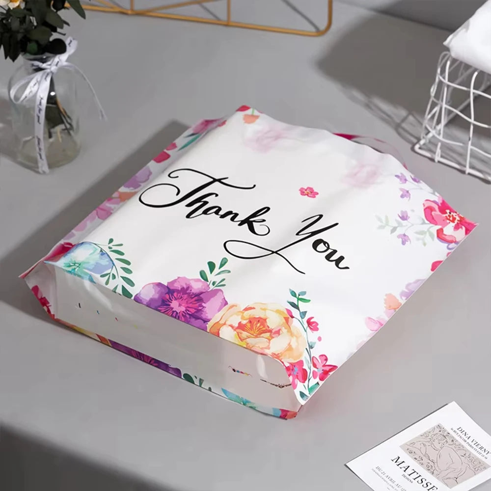 A white floral thank you gift bag with pink and green accents is displayed on a gray surface.