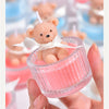A small teddy bear candle in a pink glass jar with a white ribbon being held by a hand.