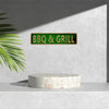 A green metal tin sign with BBQ & GRILL text mounted on a gray wall above a light stone slab.