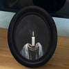 A black framed candle holder displays a lit white candle held by hands wrapped in white fabric.