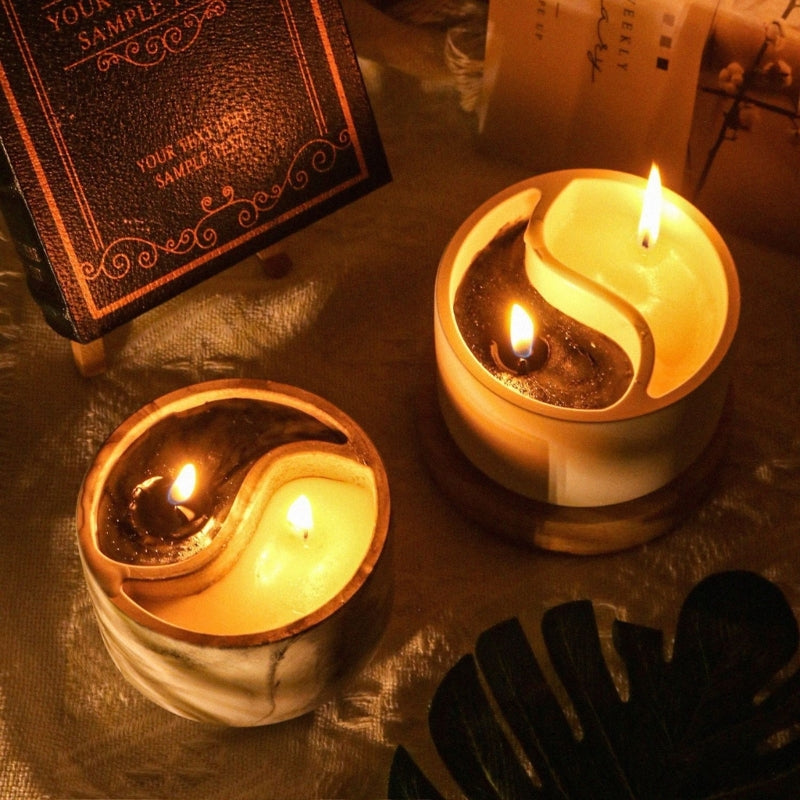 A close-up of two glowing candles in holders, set on a textured surface with a Yin Yang design.