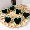 Pair of light green heart-shaped sunglasses resting on a woven mat next to a box.