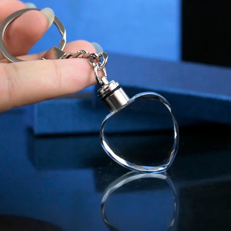 Una mano sostiene un llavero con forma de corazón de plata y un colgante de vidrio transparente y una cadena.