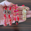 A hand holds a rosary necklace featuring red and pink beads, gold accents, and a figure of Santa Muerte.