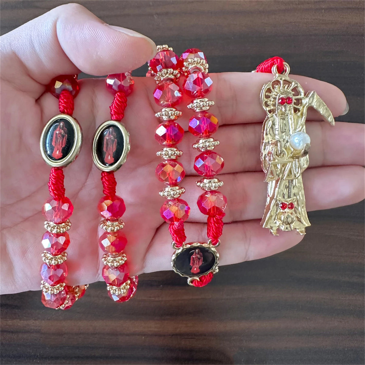 A hand holds a rosary necklace featuring red and pink beads, gold accents, and a figure of Santa Muerte.