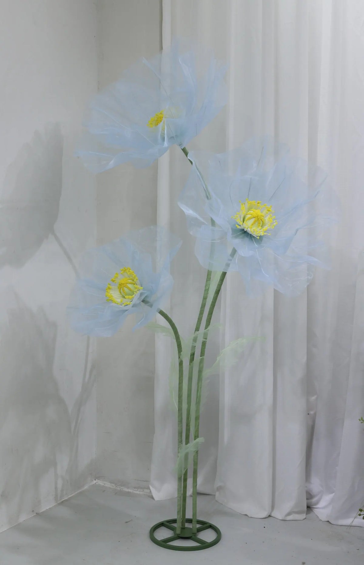 A light blue artificial silk poppy flower displayed on a green stand against a white background.