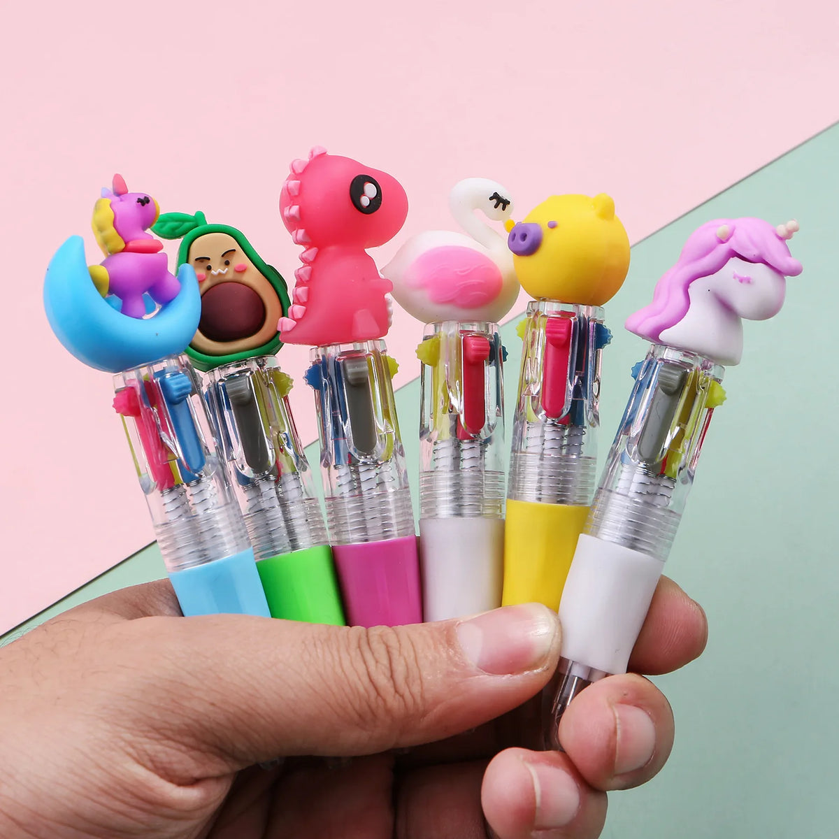 A hand holds a set of five cute cartoon mini ballpoint pens featuring characters like a pink dinosaur, a green avocado, and a