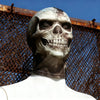A realistic 3D skull balaclava mask covering the face, worn against a rusty chain-link fence backdrop.
