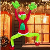 A person dressed as a green monster wearing a red sweater and bright green pants stands in front of a decorated window.