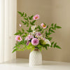 A bouquet of pink and white flowers in a white ceramic vase on a light-colored shelf.