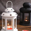 A white vintage hanging metal lantern with a glass enclosure and a lit candle sits on a wooden surface.