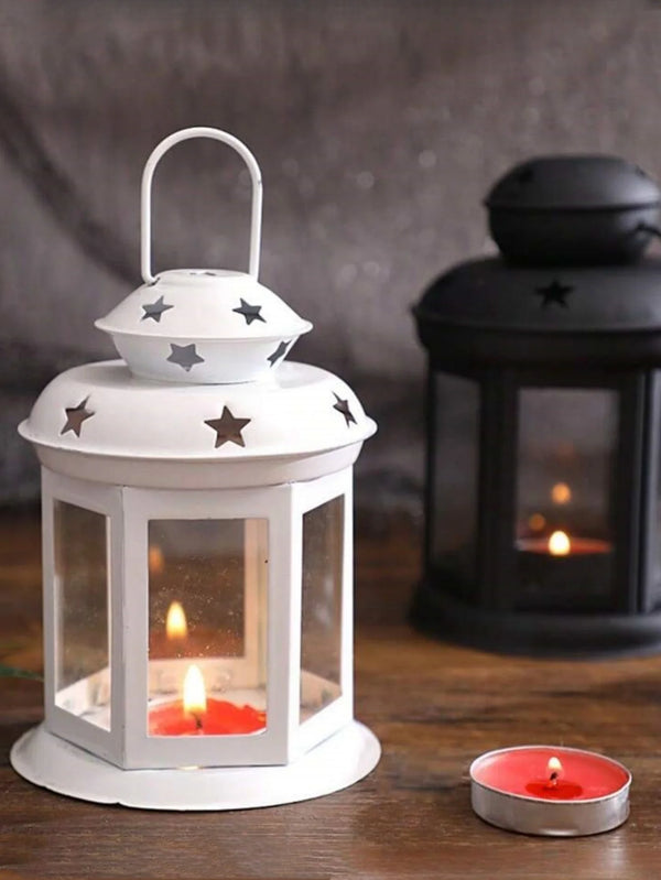 A white vintage hanging metal lantern with a glass enclosure and a lit candle sits on a wooden surface.
