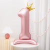 A pink foil balloon shaped like the number one stands next to a gold crown on a white table.