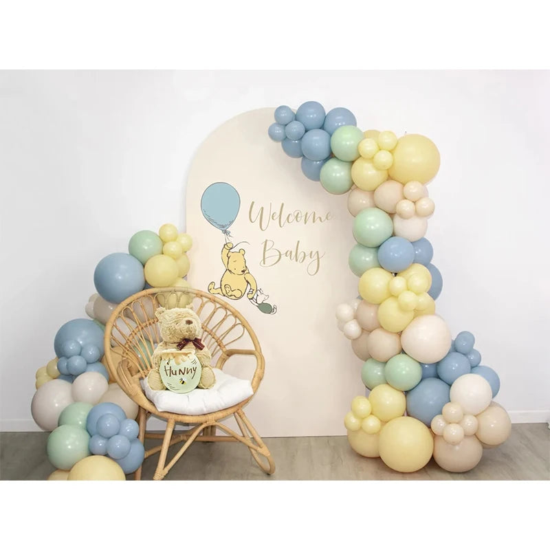 A balloon garland featuring yellow, mint green, and smoke blue balloons arranged around a wooden chair with a teddy bear.