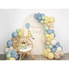 A balloon garland featuring yellow, mint green, and smoke blue balloons arranged around a wooden chair with a teddy bear.