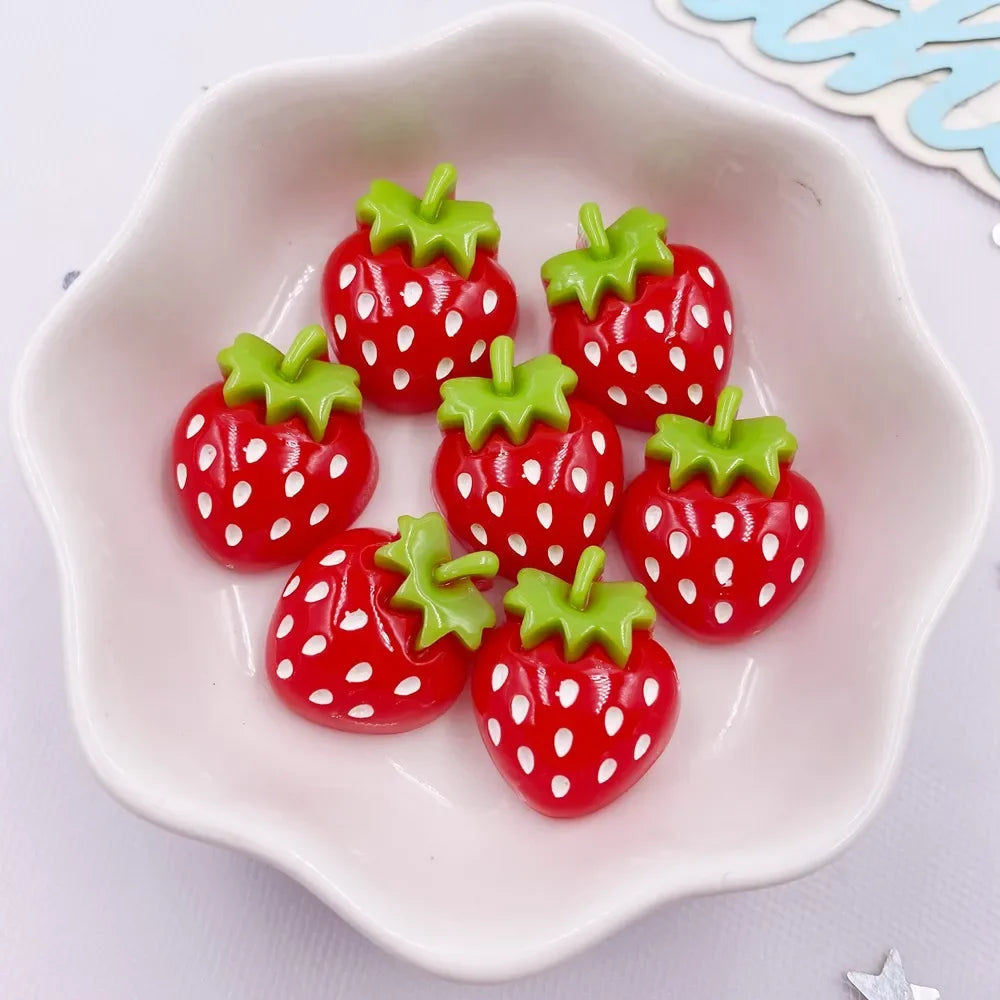 Five bright red strawberry resin flatback charms with green leaves arranged on a white ceramic dish.