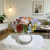 A bouquet of handcrafted blue glass flowers in a clear vase displayed on a white surface.
