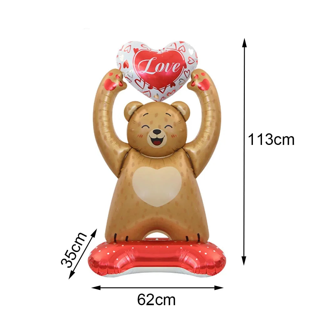 A plush bear toy with a heart-shaped balloon on its head and a heart-shaped balloon in its hands, resting on a red base.