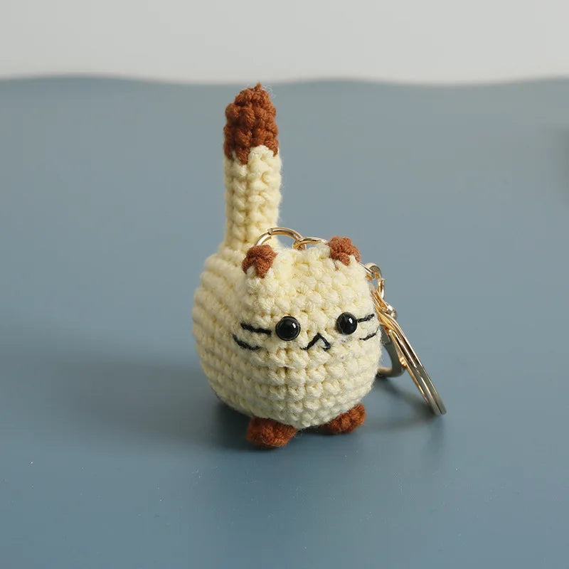 A handcrafted crochet cat keychain pendant with brown and cream yarn, resting on a gray surface.