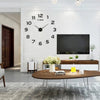 A large black acrylic wall clock with Roman numerals is mounted on a white brick wall above a wooden table.