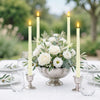 A centerpiece arrangement of white roses and greenery surrounded by tall, lit taper candles on a white table.