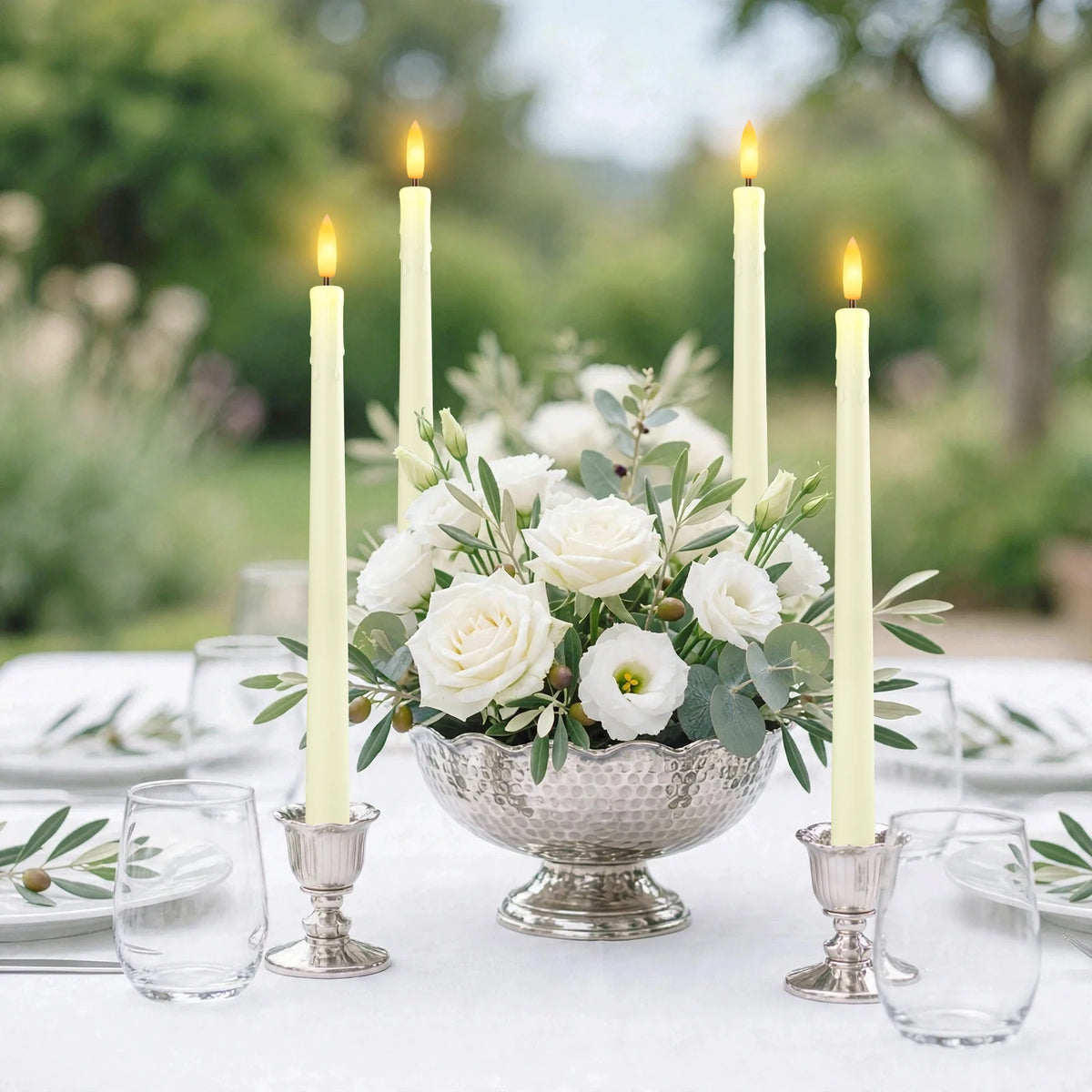 A centerpiece arrangement of white roses and greenery surrounded by tall, lit taper candles on a white table.