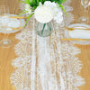 Un camino de mesa de encaje floral vintage con acentos blancos y un jarrón de flores blancas sobre una mesa de madera.