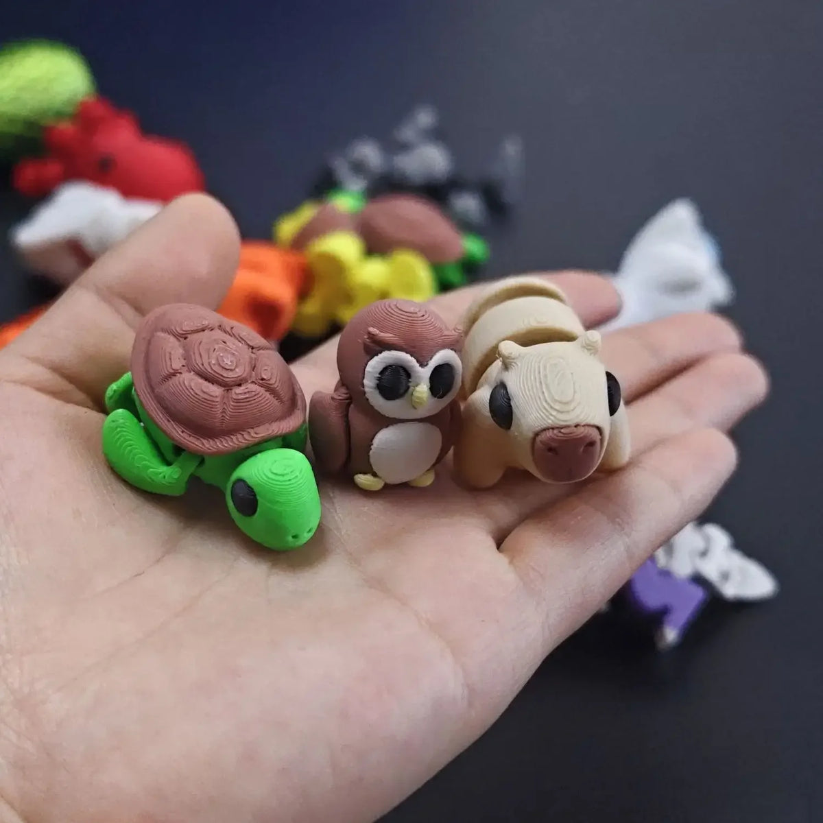 A hand holds a set of small, colorful 3D printed animal figurines including a green turtle and a brown owl.