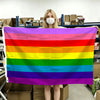 A person holds up a horizontal Pride flag banner displaying the rainbow spectrum of colors.