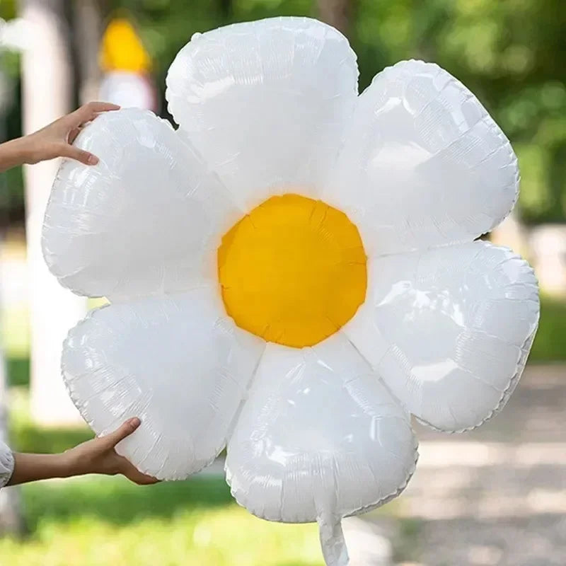 A hand is holding a white and yellow daisy-shaped aluminum foil balloon outdoors.