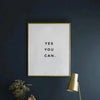 A framed canvas art print with the text YES YOU CAN on a white background mounted on a dark blue wall.