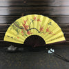 A yellow folding fan with a branch of red blossoms and birds painted on it, set against a dark wooden background.