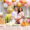 A maternity photoshoot featuring a couple and a folding dessert table display with pink and peach balloons and cupcakes.