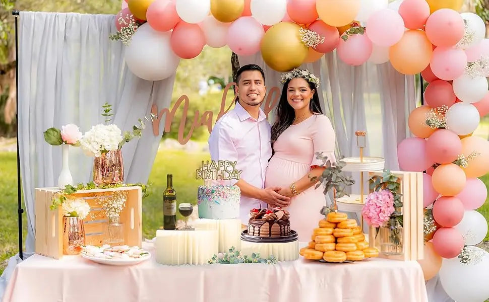 A maternity photoshoot featuring a couple and a folding dessert table display with pink and peach balloons and cupcakes.