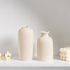 Two minimalist ceramic vases in a light beige tone displayed on a white shelf.