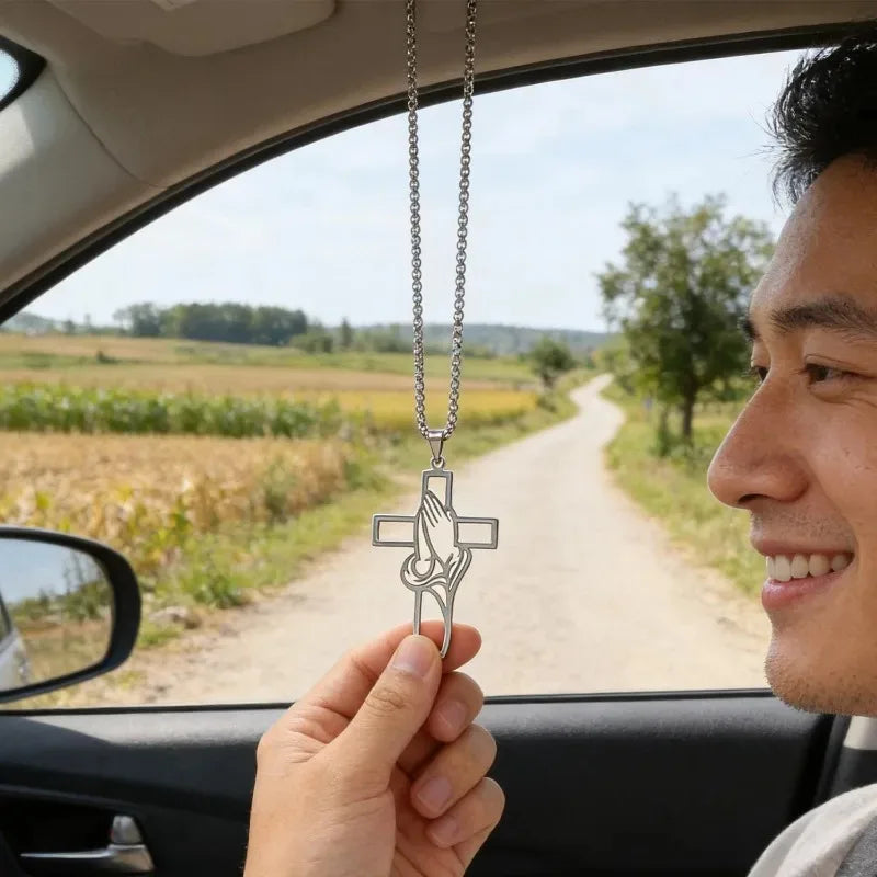 A hand holds a stainless steel praying hands cross pendant against the backdrop of a car window and rural landscape.