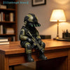 A detailed resin collectible statue of a tactical sniper soldier in olive green military gear sitting on a wooden desk.