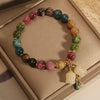 A bracelet made of various natural colored stones, including green, purple, and amber tones, displayed in a light brown box.