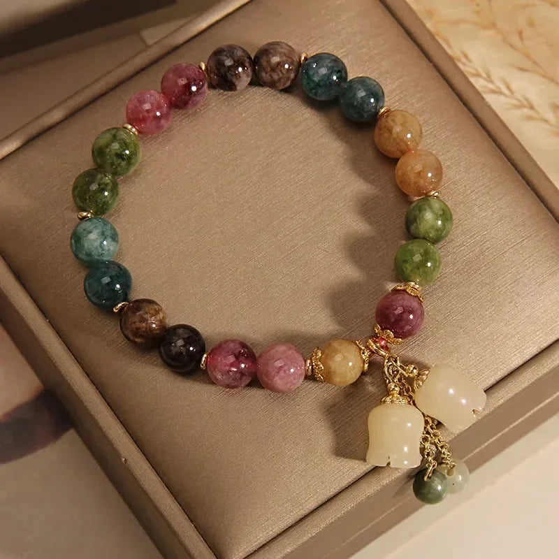 A bracelet made of various natural colored stones, including green, purple, and amber tones, displayed in a light brown box.