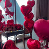 A display of red foil heart-shaped balloons and roses arranged on a dark table in an indoor setting.