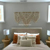 A bedroom featuring a neutral-toned bed with layered pillows and a macrame wall hanging tapestry above the headboard.