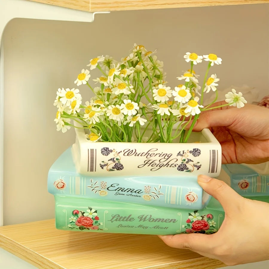 Une main tient un vase en forme de livre en céramique rempli de marguerites blanches, posé sur une pile de livres thématiques