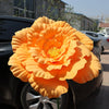 A large, vibrant orange artificial peony flower is displayed on a dark car hood.