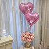 A set of pink heart-shaped foil balloons with ribbon bows displayed next to a bouquet of pink roses.