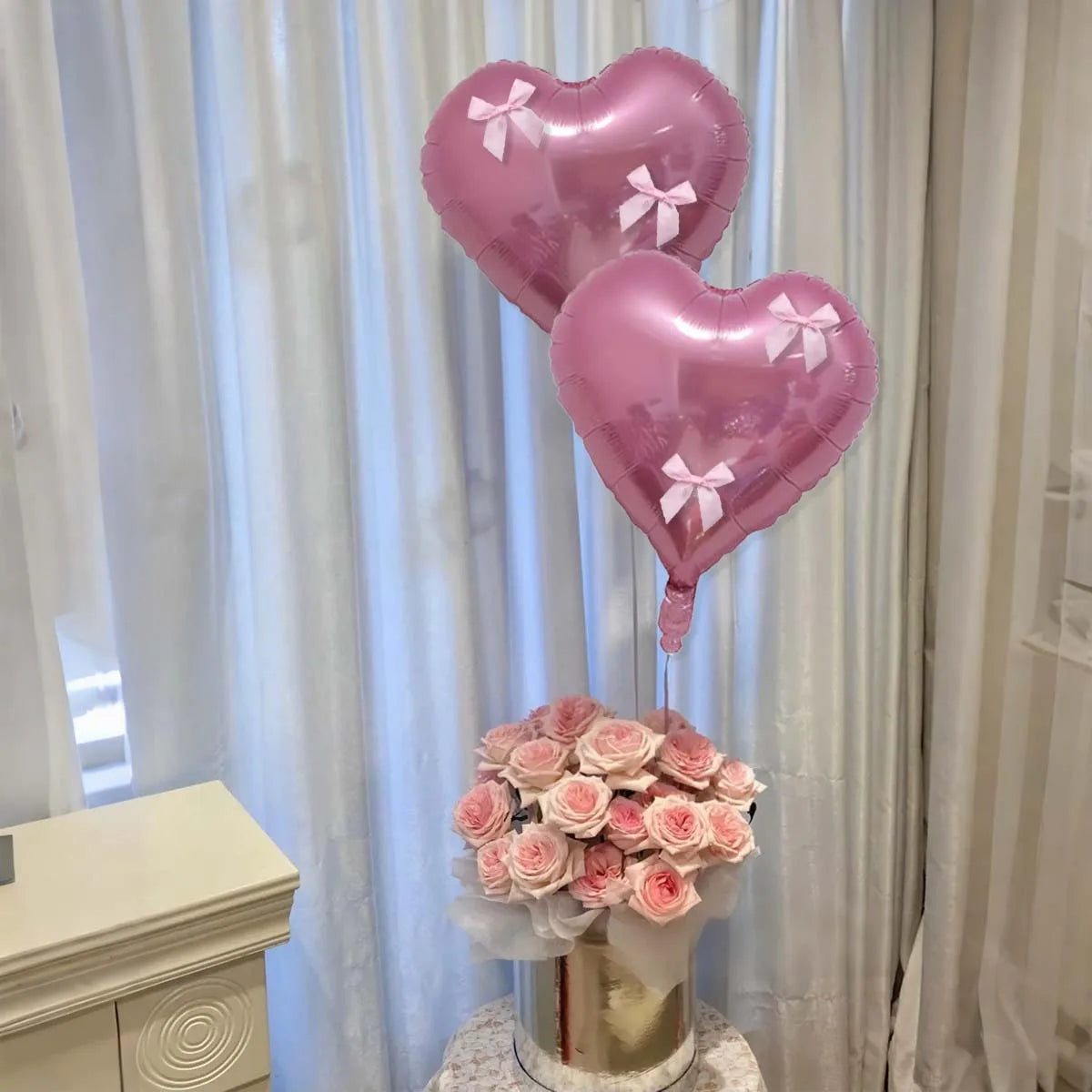 A set of pink heart-shaped foil balloons with ribbon bows displayed next to a bouquet of pink roses.
