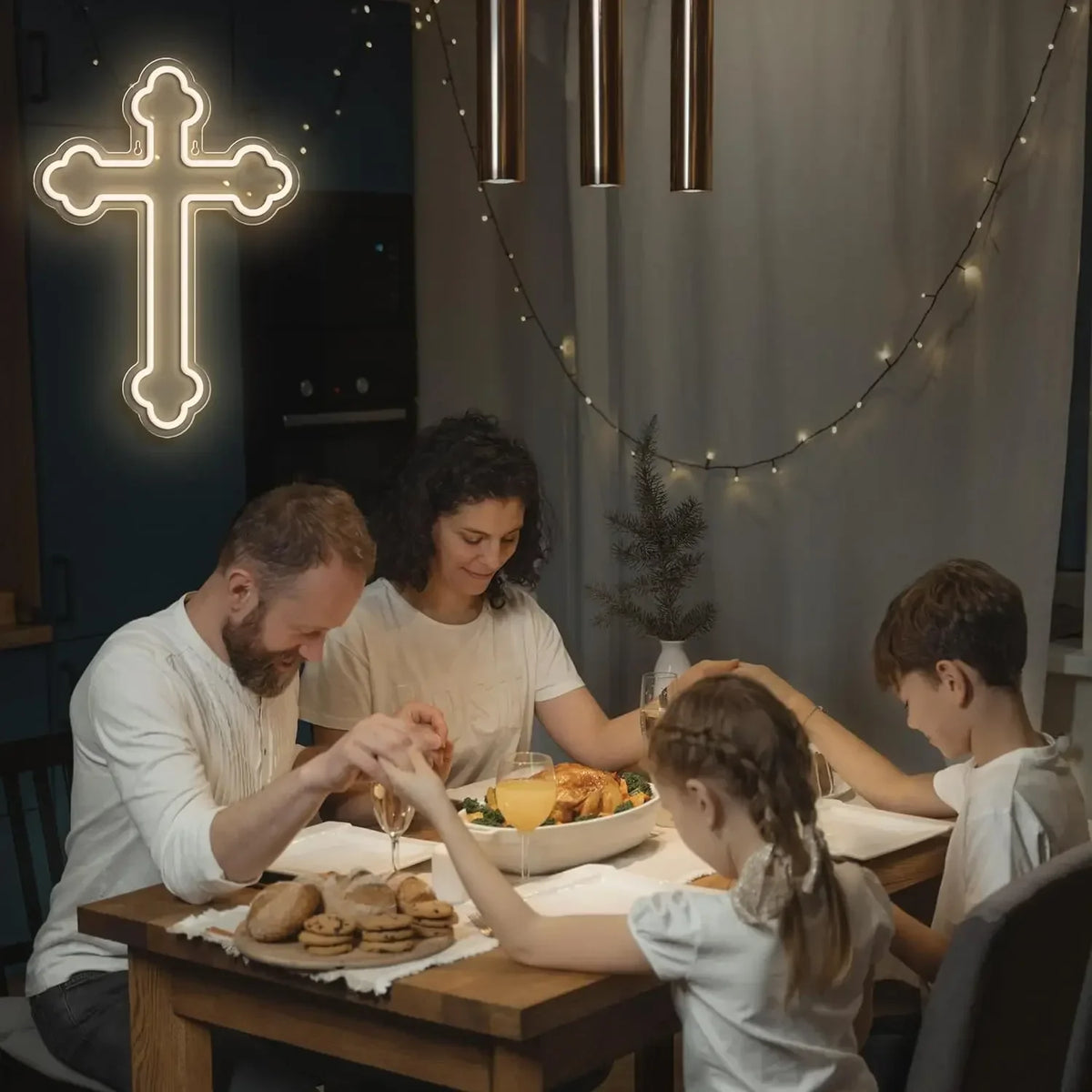 A family is gathered around a wooden table, interacting with a glowing white neon cross wall sign.